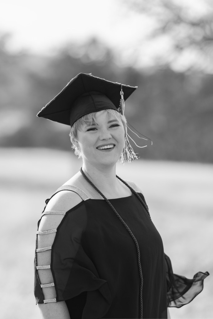 Black and white picture of Nic smiling wearing their graduate cap and cord. Link to photographer's website.